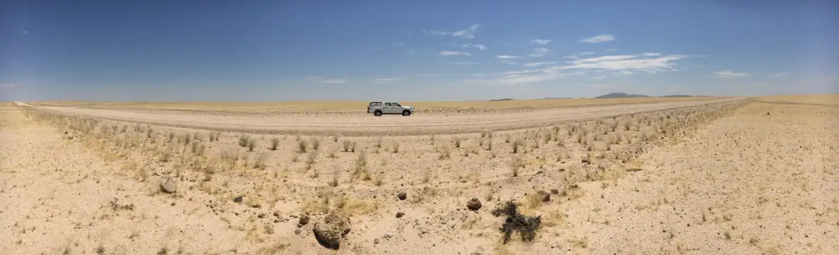 Weites Savannenland mit spärlicher Vegetation und einem weißen Geländewagen auf sandiger Piste unter blauem Himmel