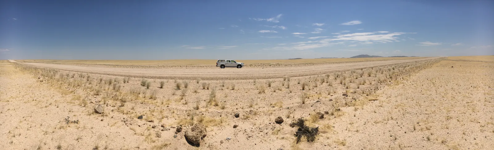 Weites Savannenland mit spärlicher Vegetation und einem weißen Geländewagen auf sandiger Piste unter blauem Himmel