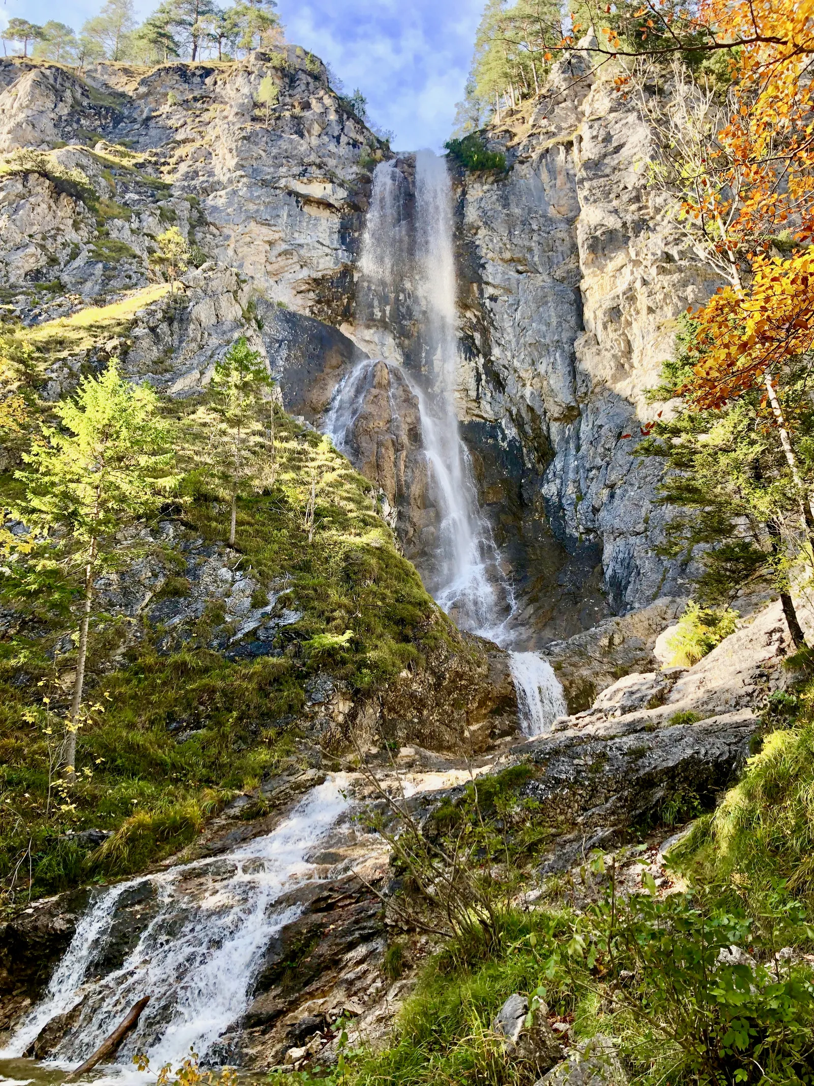 Wasserfall stürzt zwischen steilen Felswänden in einer bewaldeten Schlucht herab, unten fließt ein Bach über Felsen