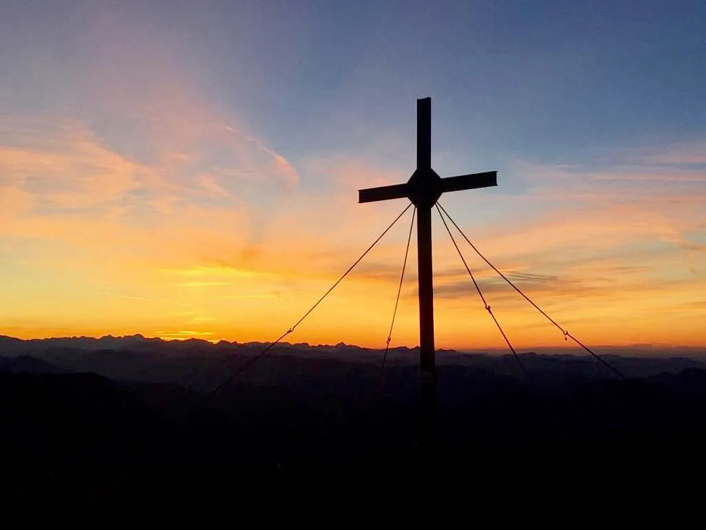 Silhouette eines Kreuzes mit Seilen auf Berggipfel vor glühendem Sonnenuntergang in Orange- und Gelbtönen