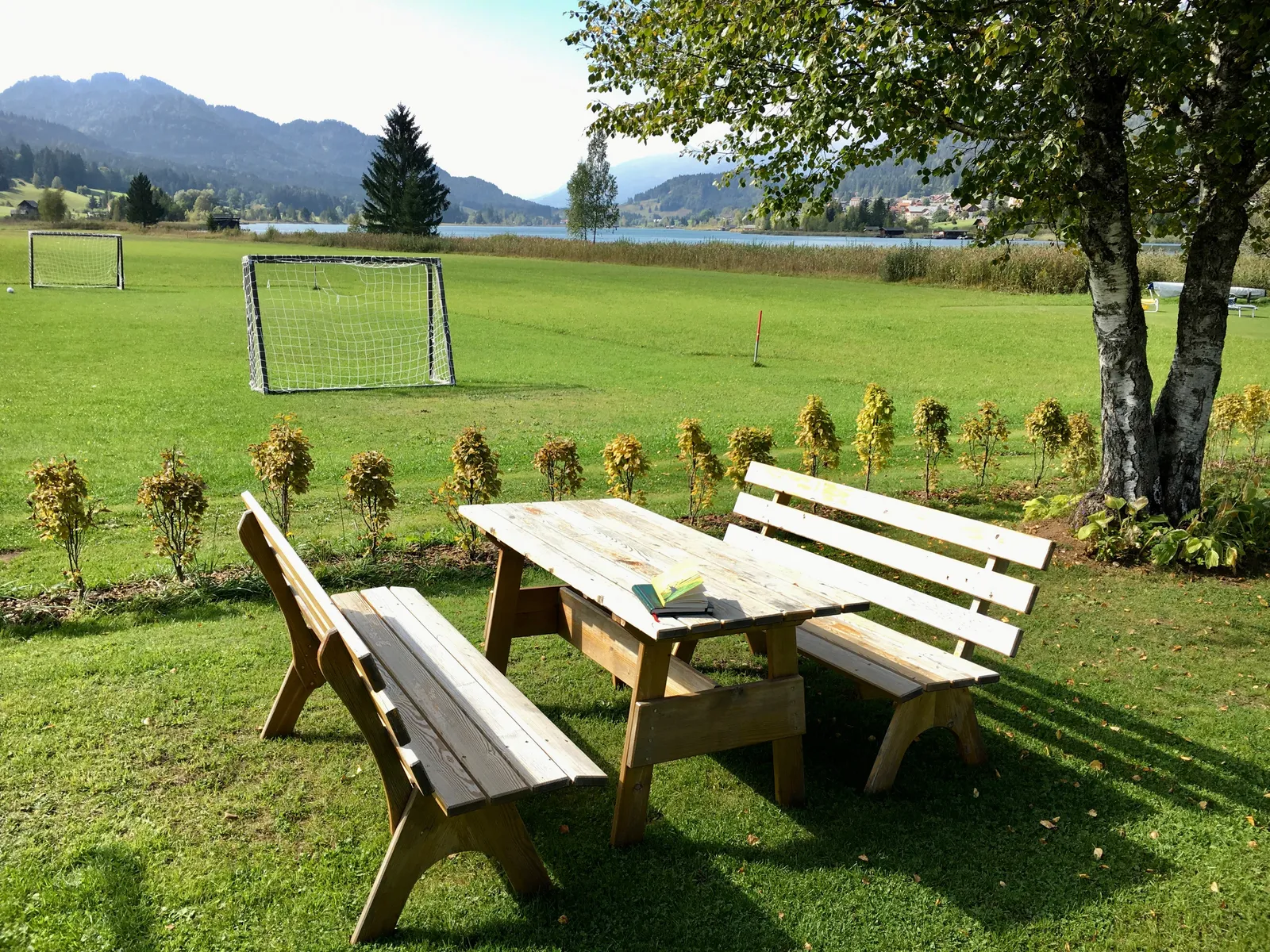 Holzpicknickbank mit Bänken auf grüner Wiese vor Bergkulisse und See, Fußballtor und junge Bäume im Hintergrund