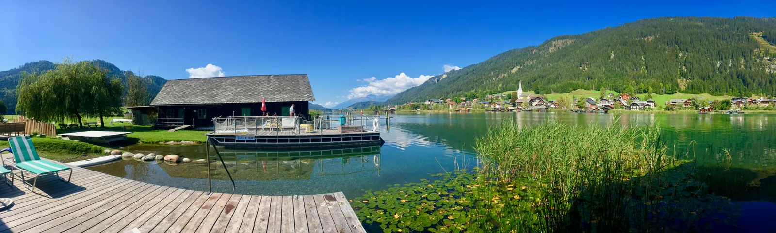 Holzsteg am türkisblauen Bergsee mit Bootshaus, Liegestuhl, Bergdorf und bewaldeten Hängen unter blauem Himmel