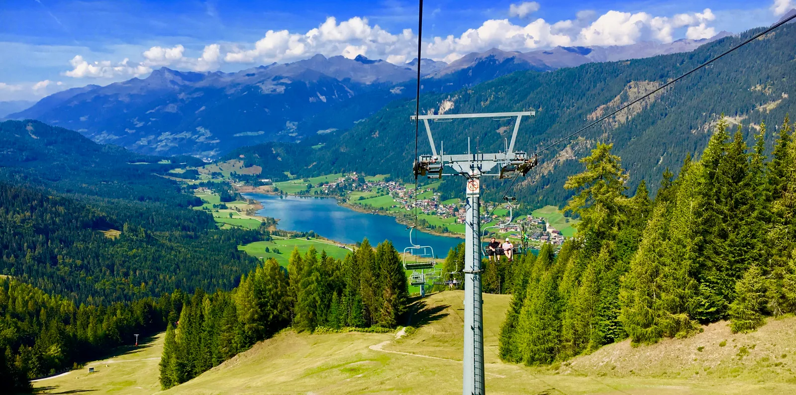 Blick vom Sessellift auf grüne Berglandschaft mit Seen, Wäldern und verschneiten Alpengipfeln unter blauem Himmel