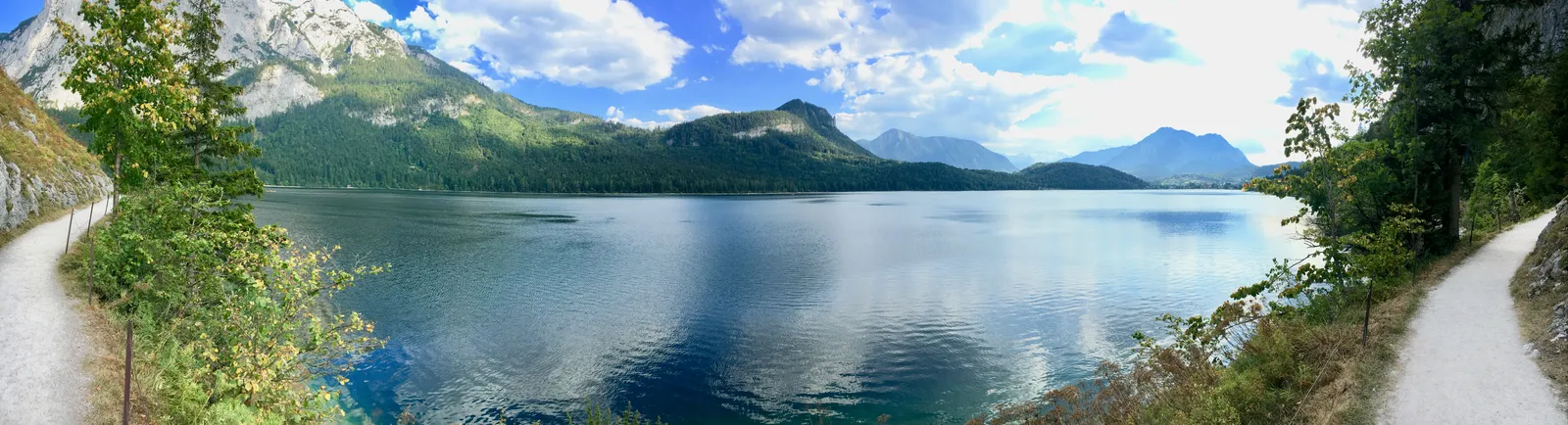 Alpensee mit bewaldeten Bergen unter blauem Himmel, von Wanderwegen mit grüner Vegetation eingefasst