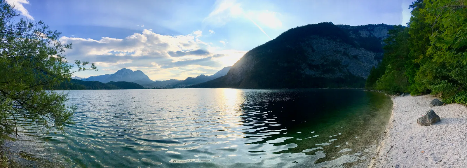 Alpiner See mit Kiesstrand, umgeben von bewaldeten Bergen unter teilweise bewölktem Himmel, spiegelnde Wasserfläche