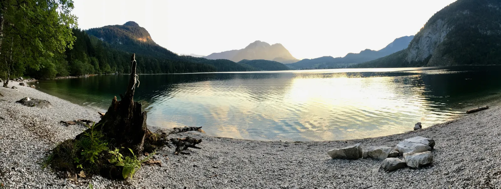 Bergseeufer mit Kiesstrand, umgeben von Nadelwald und Bergkulisse in sanftem Morgenlicht, spiegelglatte Wasseroberfläche