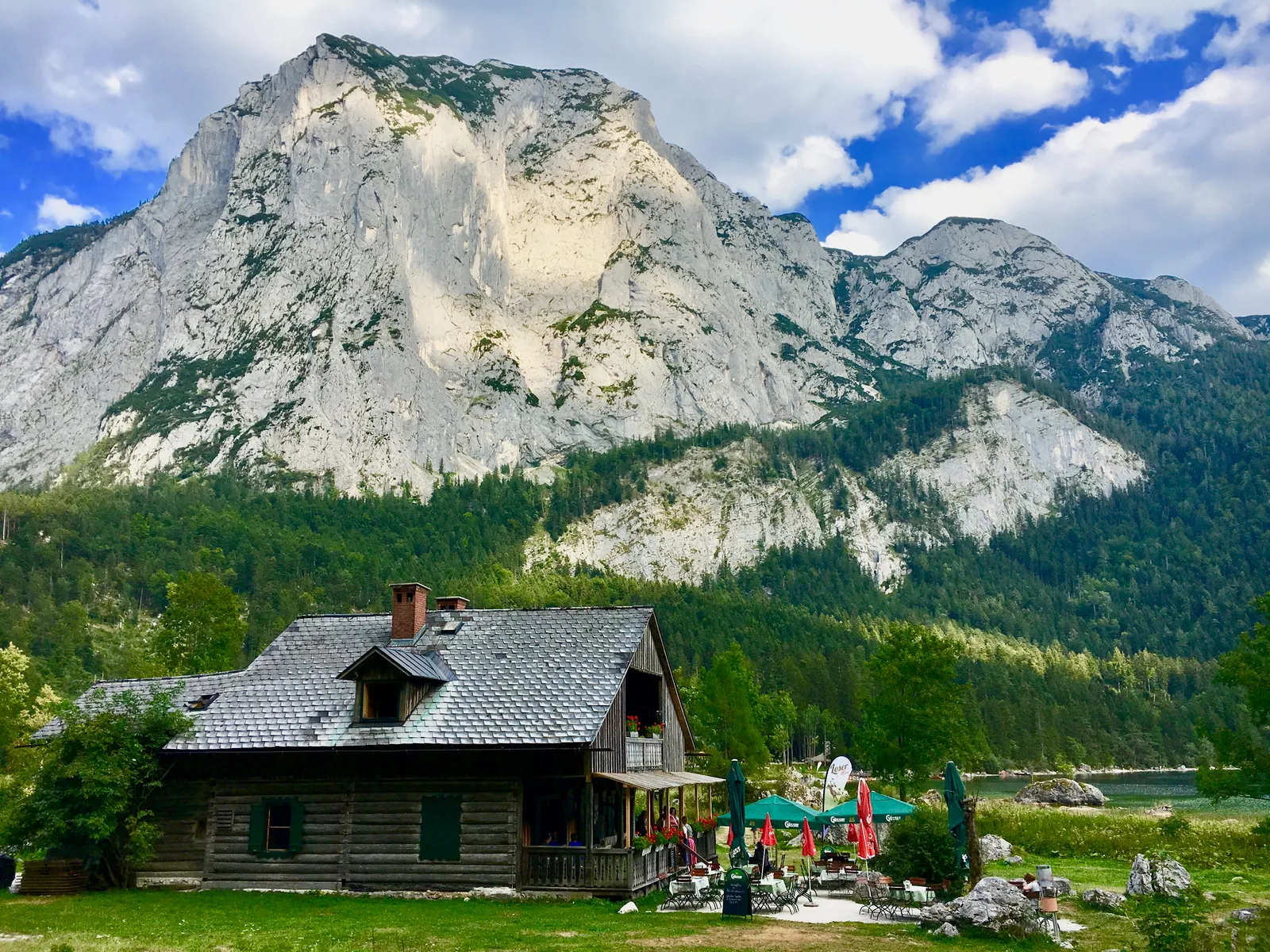 Holzhaus mit grauem Dach vor massivem Kalksteinberg, Seeufer mit Wiese, Familie spielt, klare Bergsicht