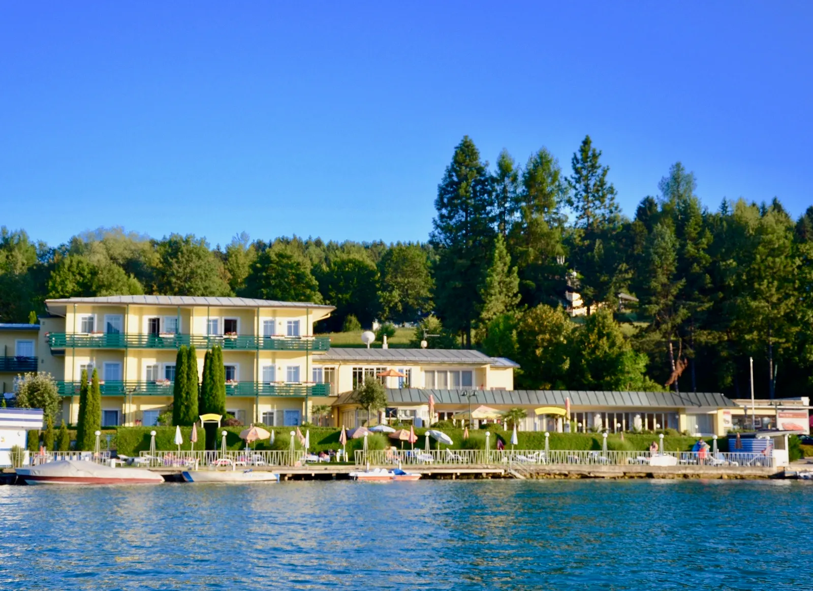 Lakefront-Hotel mit gelber Fassade, weißen Balkonen und Terrassen, dahinter bewaldete Hügel unter blauem Himmel
