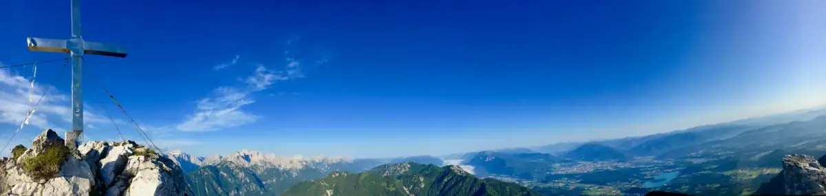 Gipfelkreuz auf Berggipfel mit weitem Blick über Bergkette und grünes Tal unter blauem Himmel