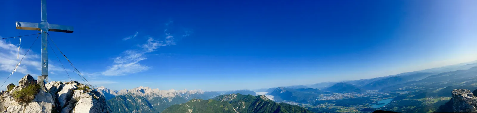 Gipfelkreuz auf Berggipfel mit weitem Blick über Bergkette und grünes Tal unter blauem Himmel