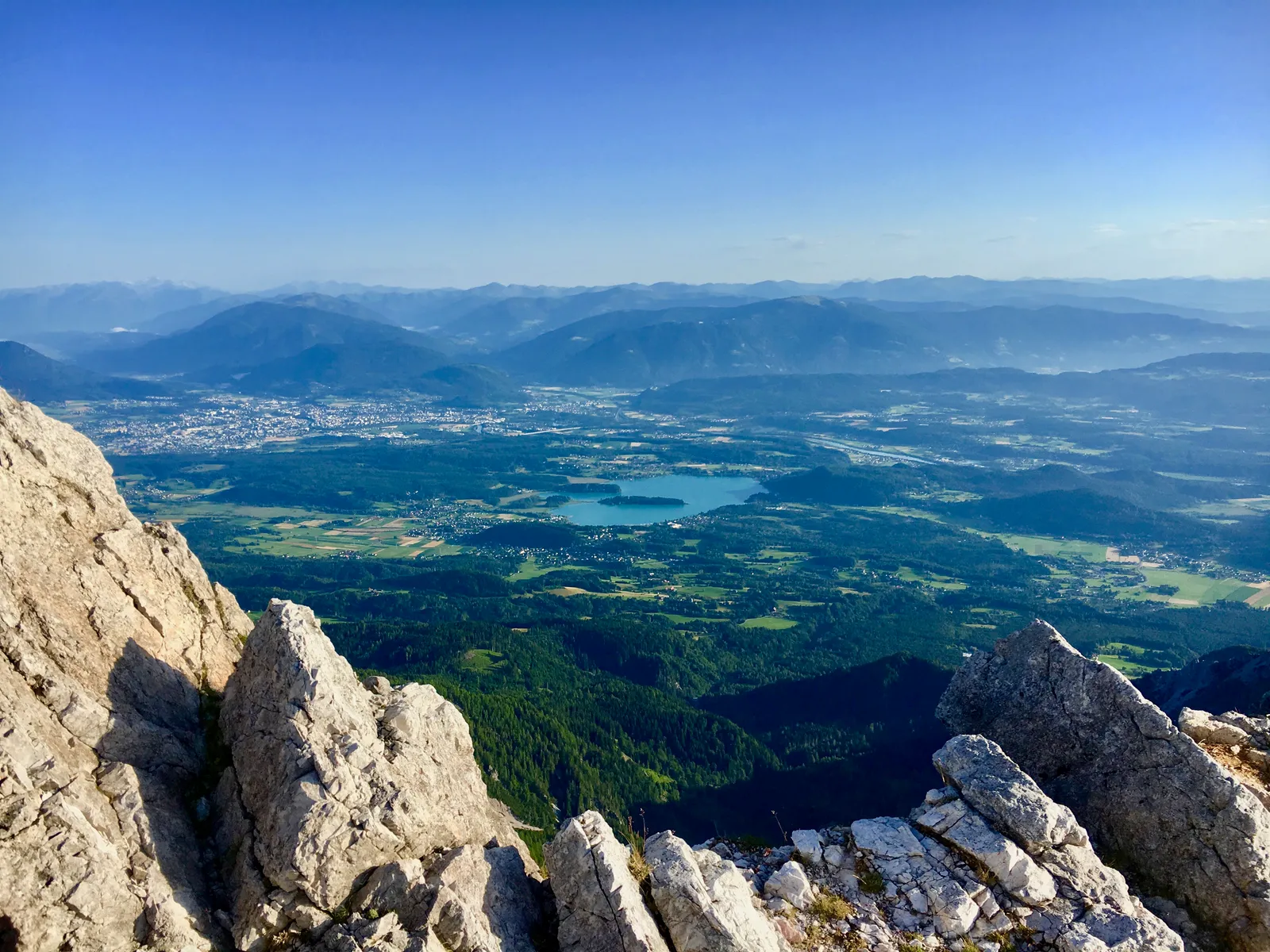 Blick vom Mittagskogel über Bergketten auf grüne Täler, Seen und Siedlungen im weiten Alpenvorland bei klarem Wetter