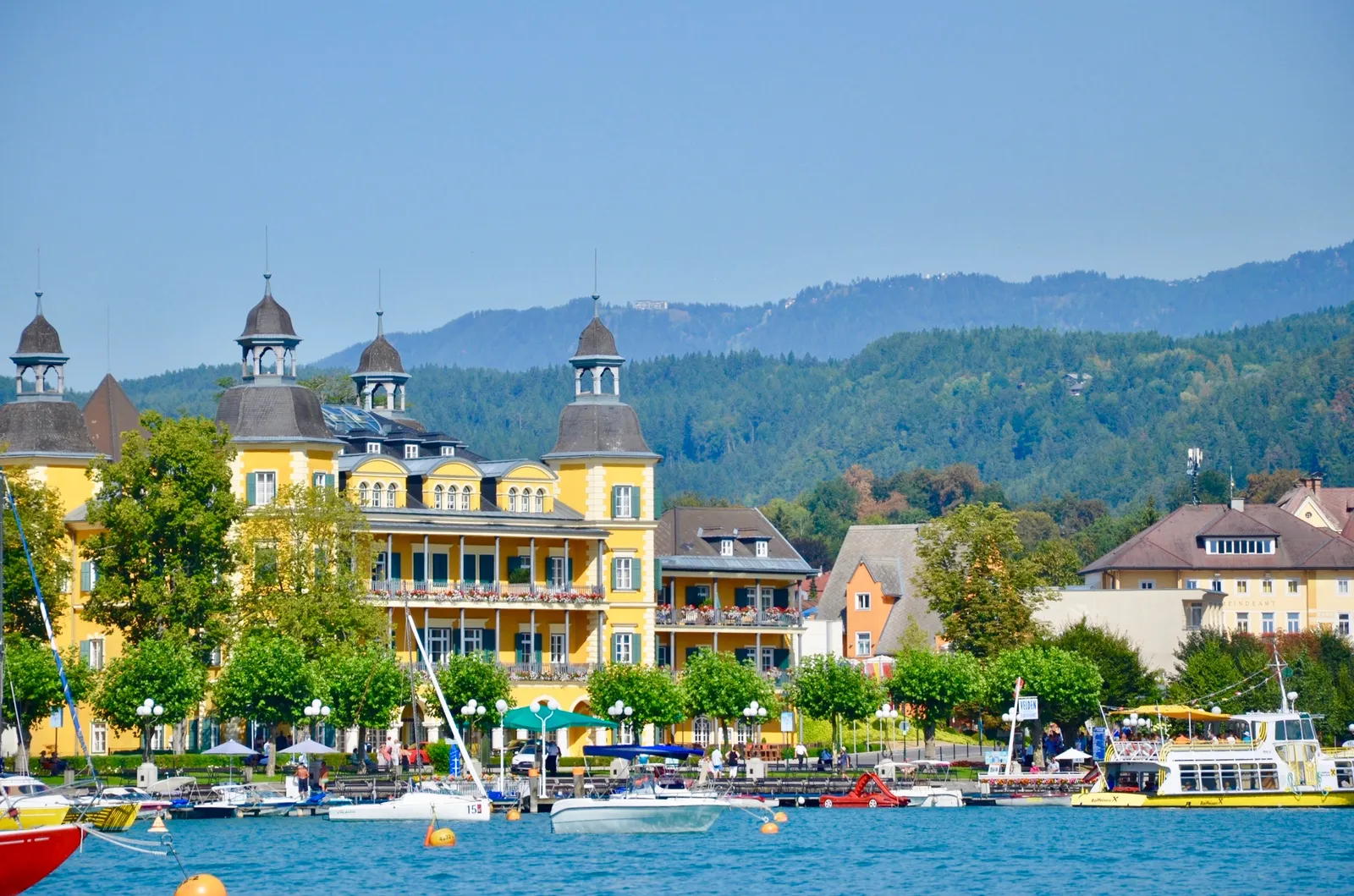 Gelbes Schloss am Ufer mit Booten, dahinter bewaldete Berge und klares blaues Wasser unter sonnigem Himmel