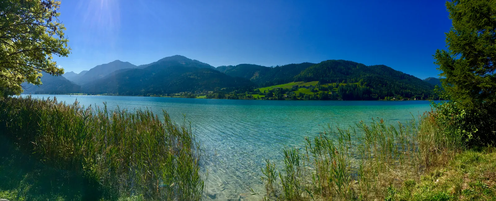 Klare türkisgrüne Seefläche mit Schilfgürtel im Vordergrund, dahinter bewaldete Bergkette unter blauem Himmel