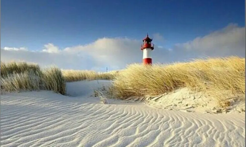 Leuchtturm mit rot-weißem Anstrich hinter Sanddünen mit Strandhafer und flacher Wasserfläche unter blauem Himmel