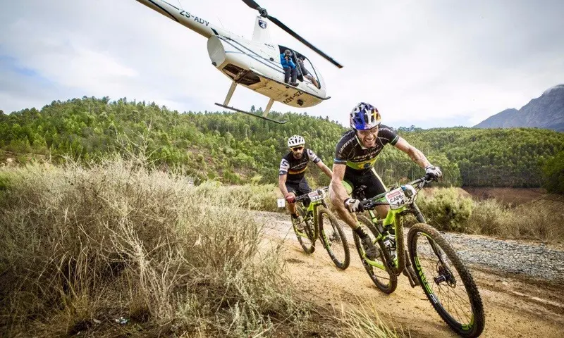 Zwei Mountainbiker fahren auf sandiger Strecke durch buschige Landschaft, während ein Hubschrauber über ihnen schwebt