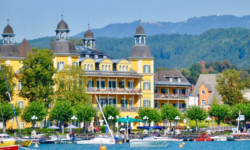 Gelbes Schloss mit Türmen am Seeufer, davor Boote und Badegäste, bewaldete Berge im Hintergrund