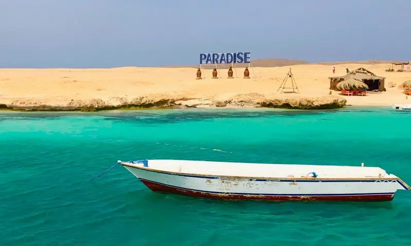 Türkisblaues Wasser mit vertäutem Fischerboot vor Sandstrand, "Paradise"-Schriftzug