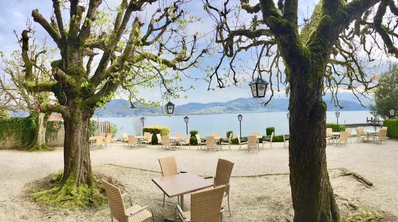 Gemütlicher Schanigarten am See mit Tischen unter blühenden Bäumen, ruhige Frühjahrsatmosphäre mit Bergblick
