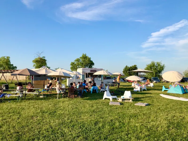 Sommerliches Treffen an einem See mit Menschen unter Sonnenschirmen, Camper und Zelten auf grüner Wiese unter blauem Himmel
