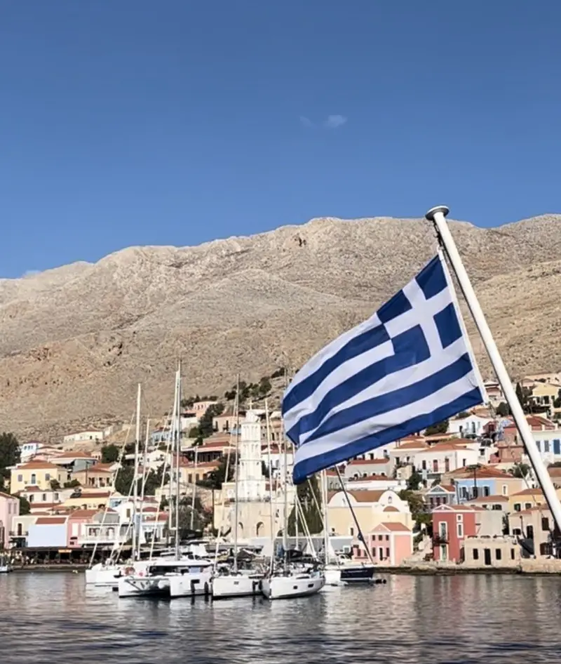 Griechische Flagge weht an Segelboot im Hafen, bunte Häuser und kahle Berge im Hintergrund, klarer Himmel