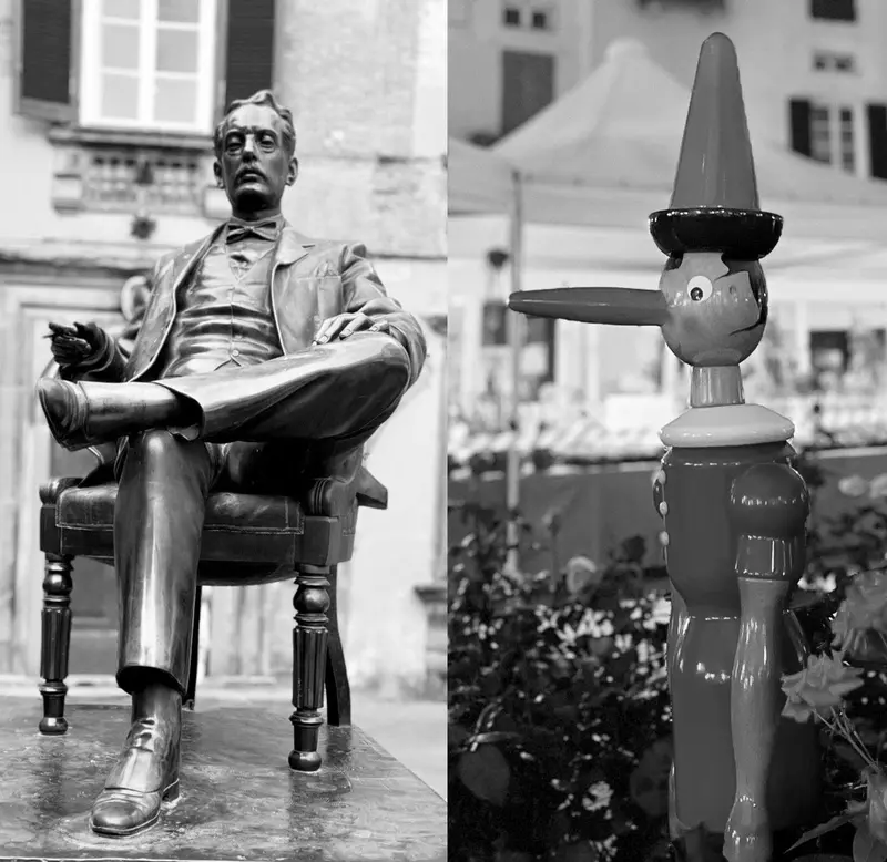 Statue eines sitzenden Mannes mit Zeitung und bunte Vogelfigur in mediterraner Stadtkulisse, Schwarz-Weiß-Fotografie