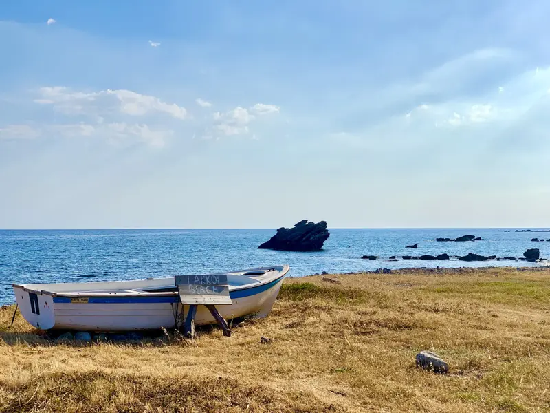 Zwei Fischerboote liegen auf steinigem Strand, dahinter ruhiges Meer mit Felsen und blauem Himmel