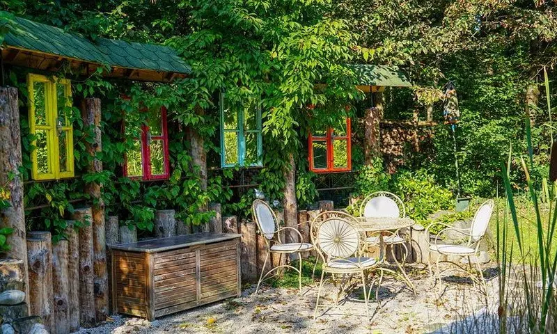 Verwunschener Garten mit efeuberankem Häuschen, bunten Fensterrahmen und Gartenmöbeln auf Kiesfläche