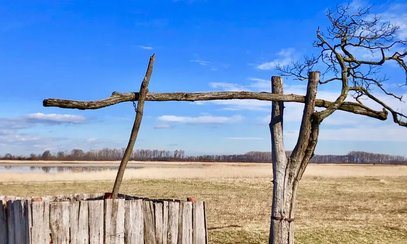 Alte Holztor-Konstruktion in flacher Landschaft mit ausgetrocknetem Boden und Bäumen am Horizont unter blauem Himmel