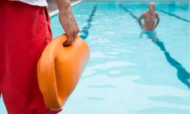Rettungsschwimmer in roter Badekleidung hält orangefarbenes Rettungsgerät, im Hintergrund Badegäste im Schwimmbecken