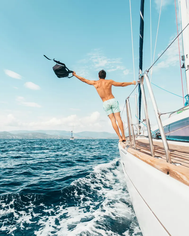 Mann springt von Segelboot ins Meer, hält Schnorchelausrüstung in der Hand, klares Wasser und Berglandschaft im Hintergrund