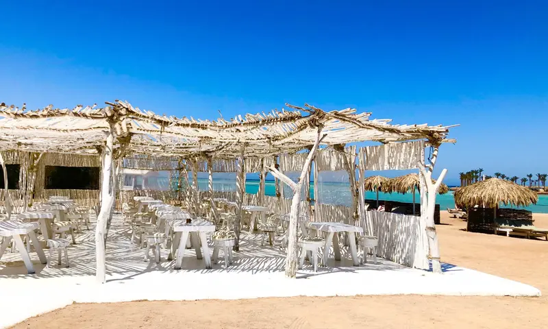 Strandbar mit weißen Holzstrukturen und Strohdächern an einem sonnigen Strand mit türkisblauem Meer