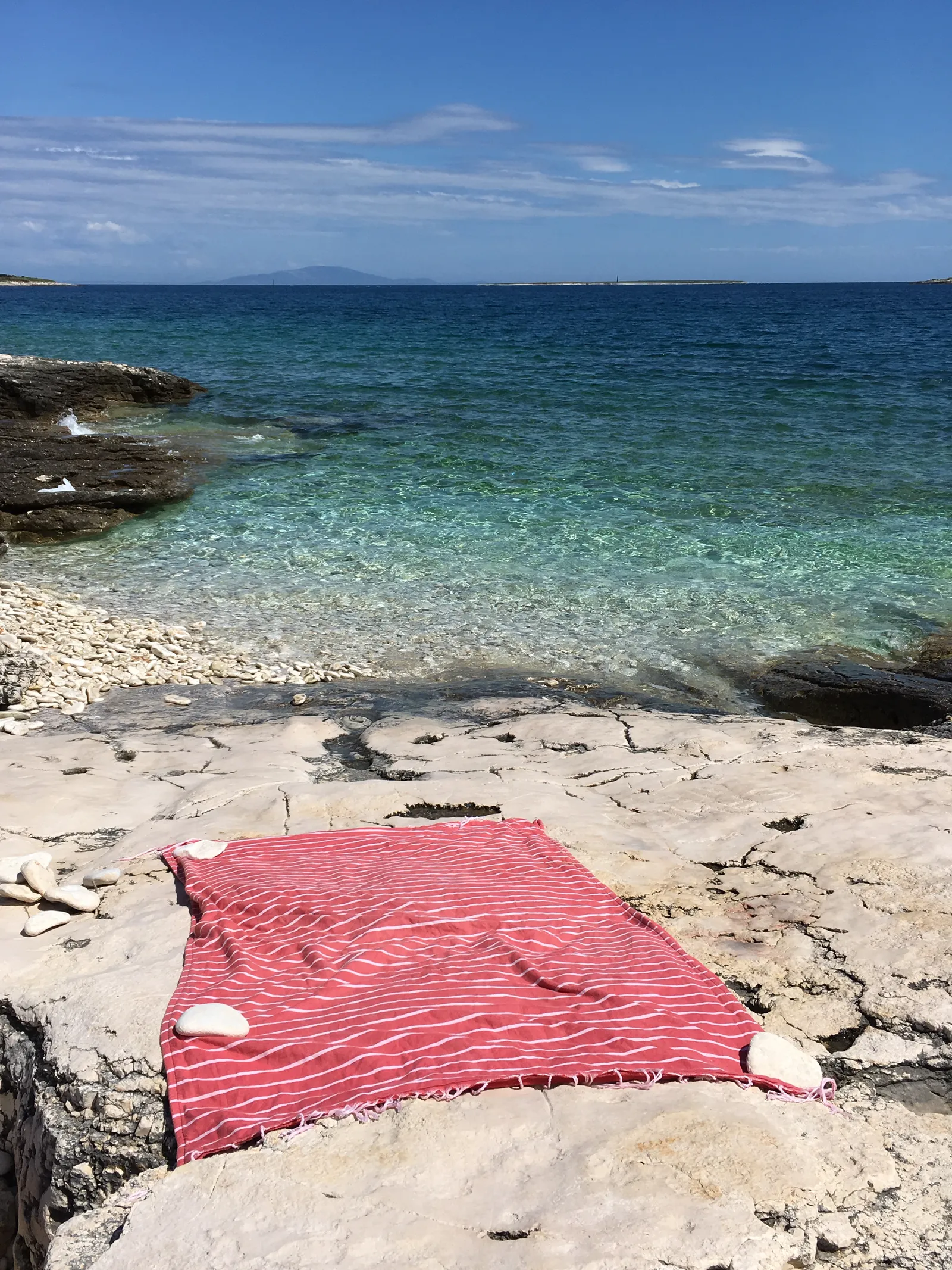 Rotes Strandtuch auf Kiesstrand mit kristallklarem Meerwasser und felsenküste unter blauem himmel