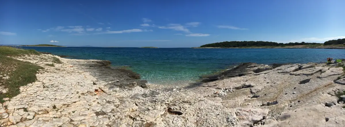 Steiniger Strand mit kristallklarem Wasser, bewaldeter Hügellandschaft