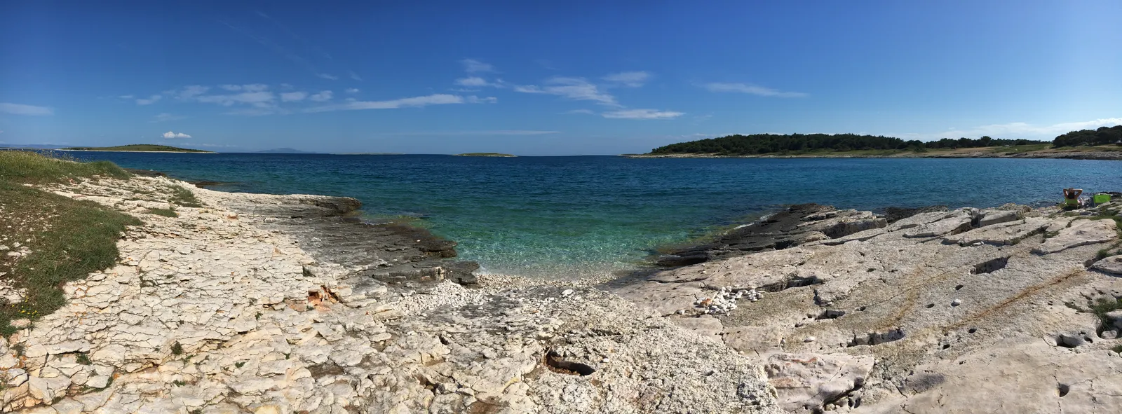 Steiniger Strand mit kristallklarem Wasser, bewaldeter Hügellandschaft