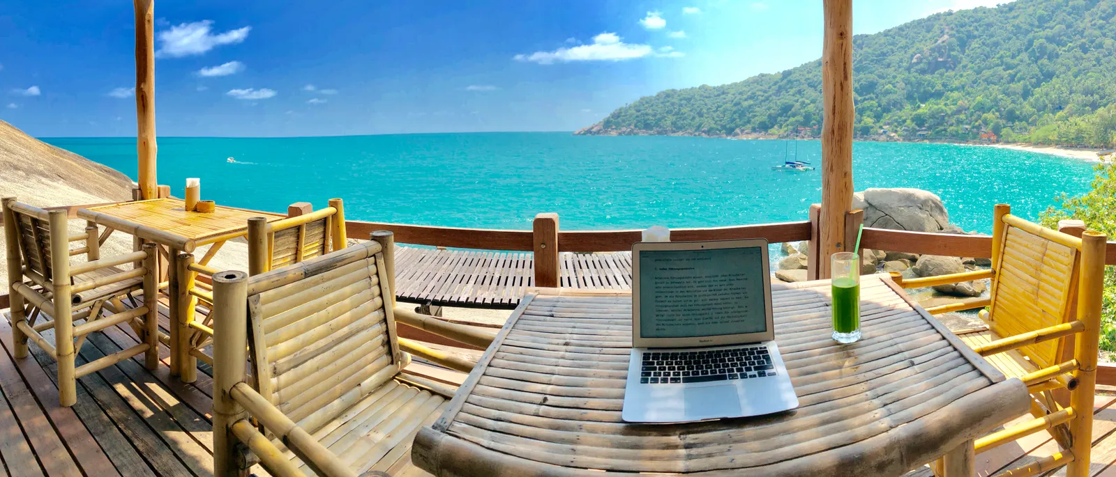 Holzterrasse mit Laptop und Getränk am türkisfarbenen Meer, Bucht mit bewaldeten Hügeln und Booten im Hintergrund