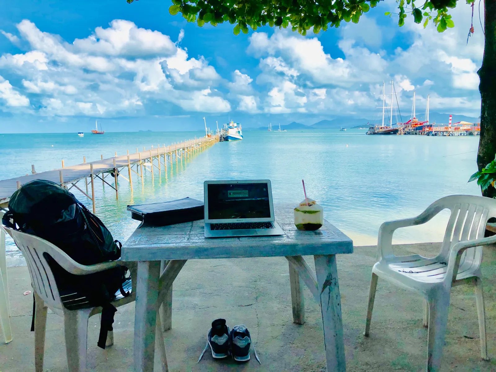 Arbeitsplatz am Strand mit Laptop auf blauem Tisch, weiße Stühle, türkisfarbenes Meer, Holzsteg und Segelboote im Hintergrund