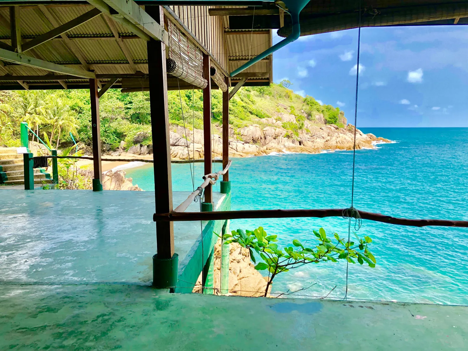 Yoga-Plattform mit Holzunterstand an tropischer Bucht, türkises Wasser, grüne Hügel und Felsen, klarer Himmel