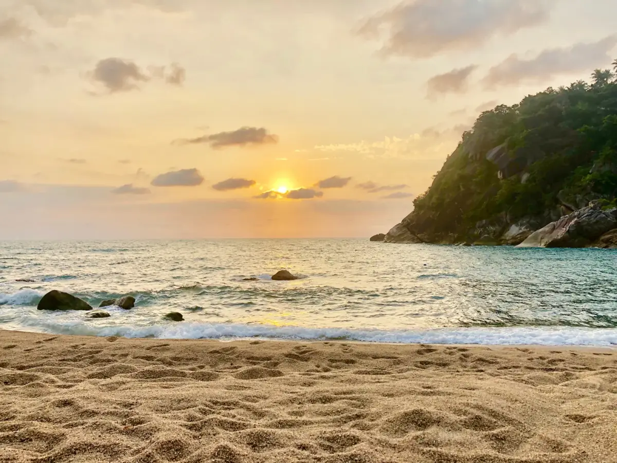 Tropische Bucht bei Sonnenaufgang mit Sandstrand, Wellen, Felsen im Wasser und bewaldeter Klippe am Horizont