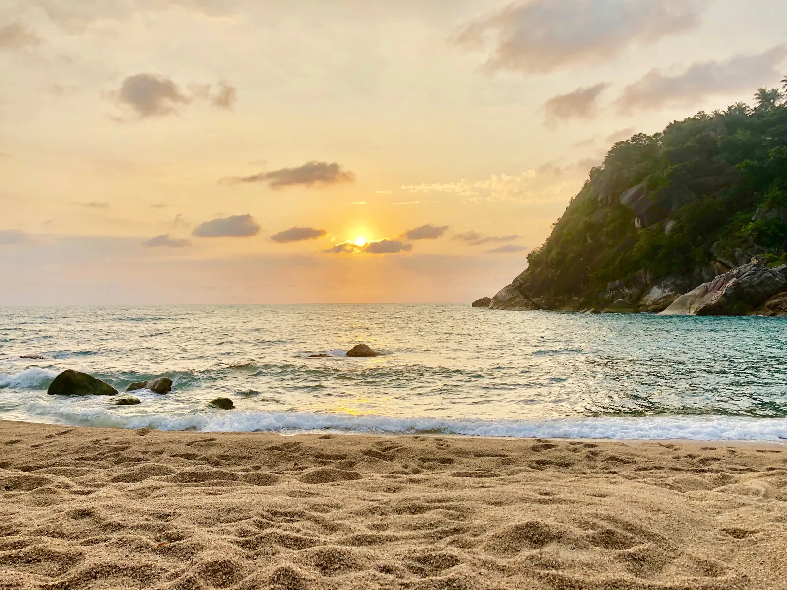 Tropische Bucht bei Sonnenaufgang mit Sandstrand, Wellen, Felsen im Wasser und bewaldeter Klippe am Horizont