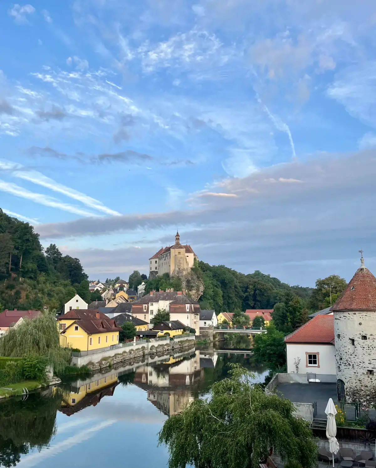Flusslandschaft mit Burg auf Hügel, gelben und weißen Häusern, spiegelndem Wasser und dramatischem Himmel bei Sonnenuntergang