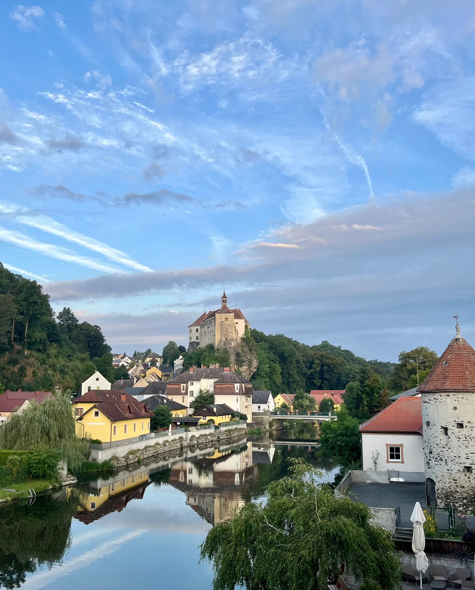 Flusslandschaft mit Burg auf Hügel, gelben und weißen Häusern, spiegelndem Wasser und dramatischem Himmel bei Sonnenuntergang