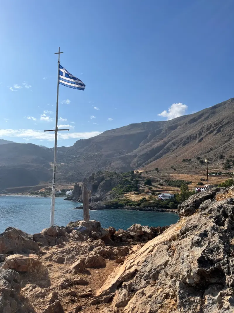 Griechische Flagge an weißem Holzkreuz auf felsiger Küste mit türkisblauem Meer