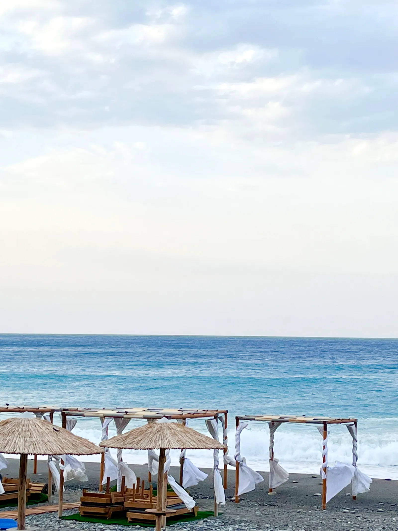 Strandliegen mit Strohdächern und weißen Tüchern am schwarzen Kiesstrand, dahinter türkisfarbenes Meer und bewölkter Himmel