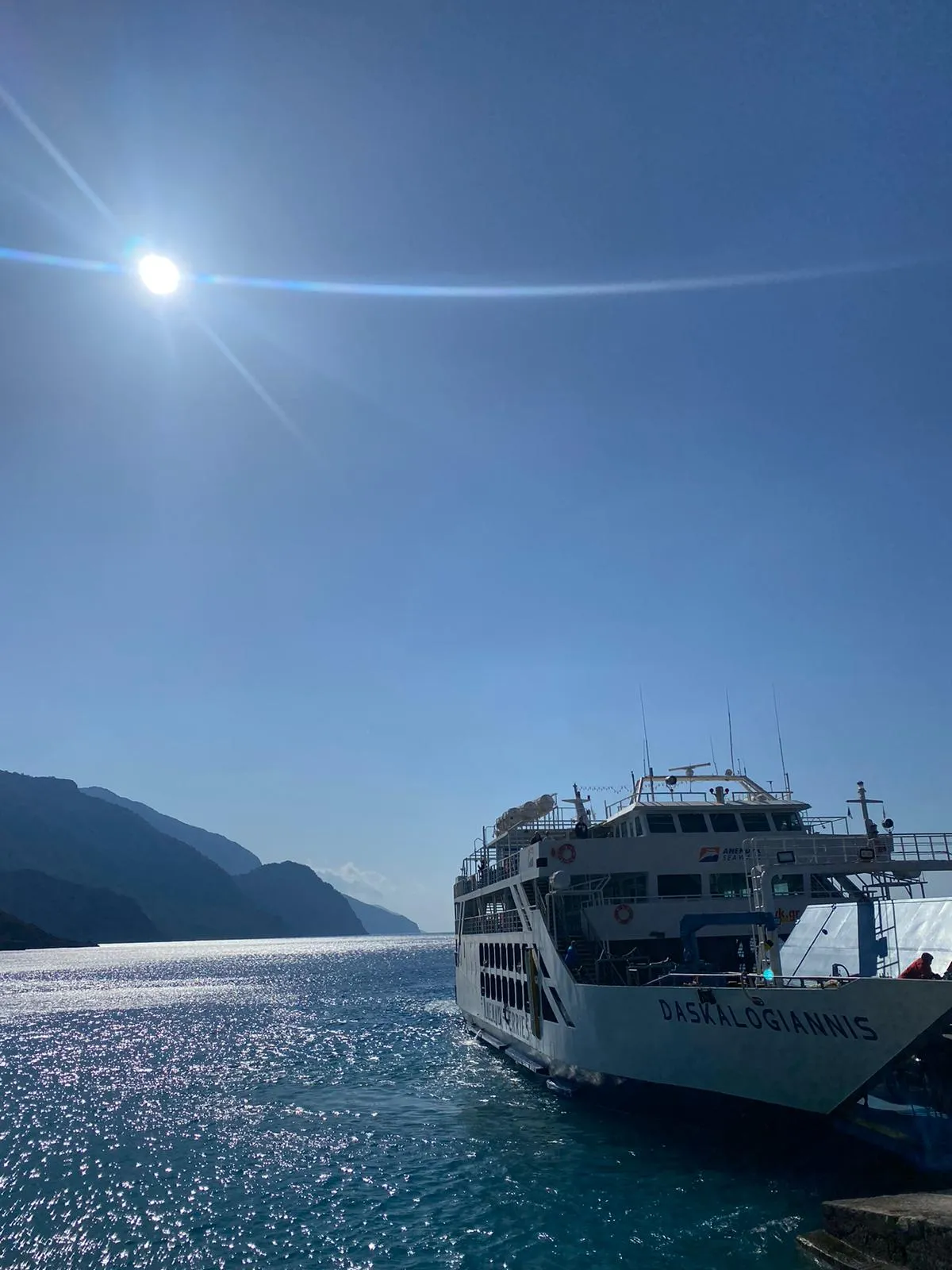 Fährschiff Daskalogiannis auf türkisblauem Meer vor Bergkulisse, Passagiere an Deck bei Sonnenschein