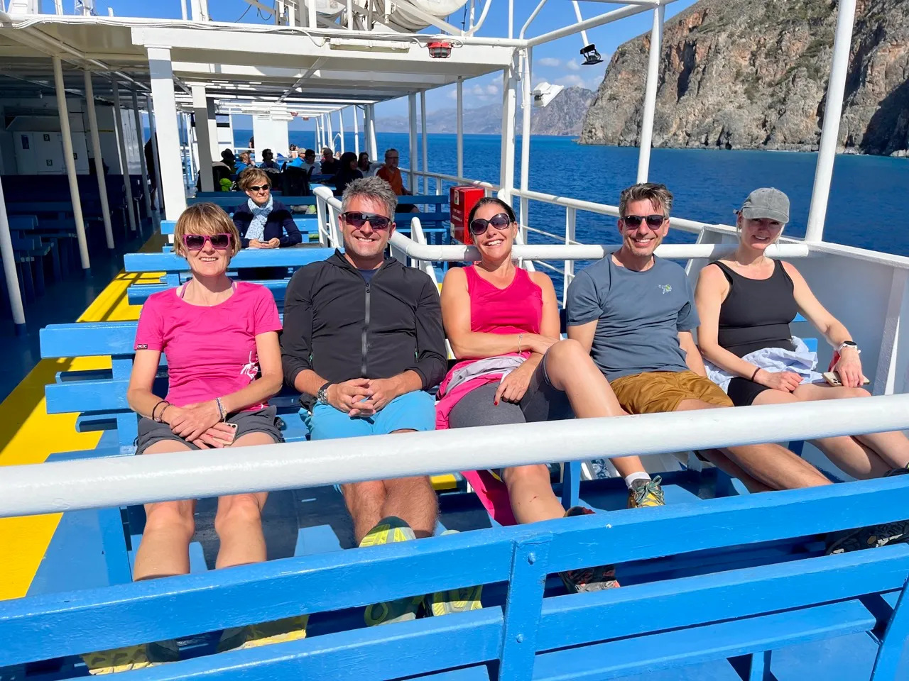 Fünf Personen sitzen auf einer blauen Fährbank mit Blick auf türkisblaues Meer und imposante Felsklippen an der Südküste