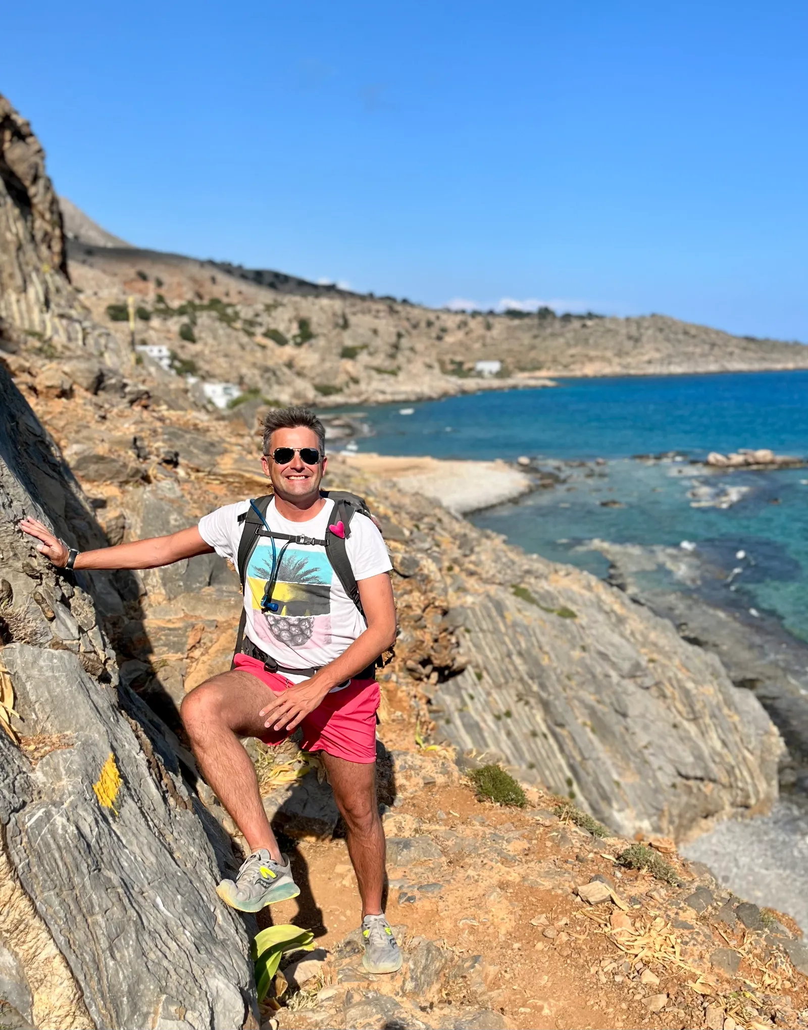 Mann mit Rucksack posiert an Steilküste über türkisfarbenem Meer mit Strand und Siedlung, Wanderung Südkreta