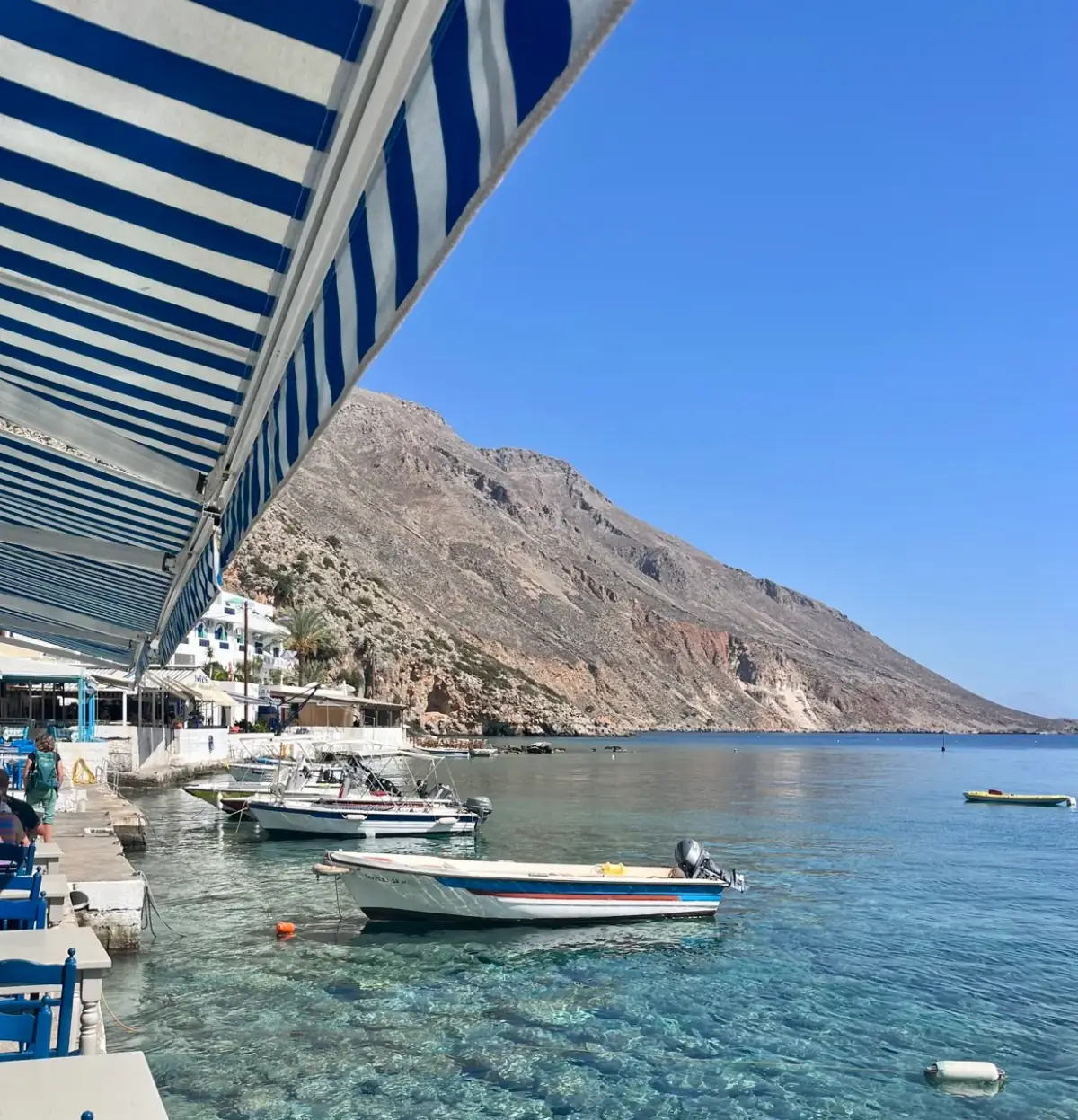 Fischerboote liegen in kristallklarem Wasser vor steilen Felsklippen, während ein blauweißer Sonnenschirm Schatten spendet