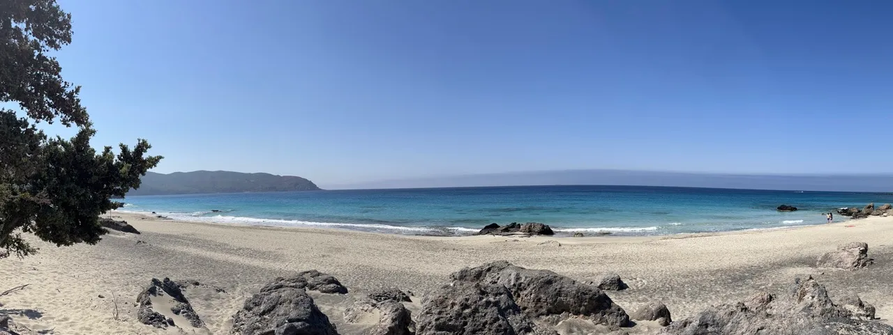 Breiter Sandstrand mit türkisblauem Meer, felsigen Buchten und Bergen im Hintergrund unter klarem Himmel
