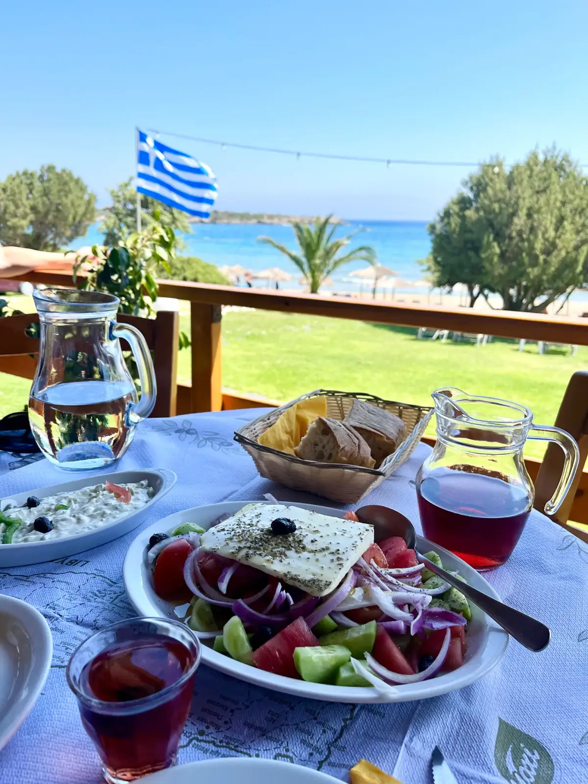 Festliche Mahlzeit mit Feta-Salat und Rotwein auf Terrasse mit Meerblick, griechische Flagge im Hintergrund