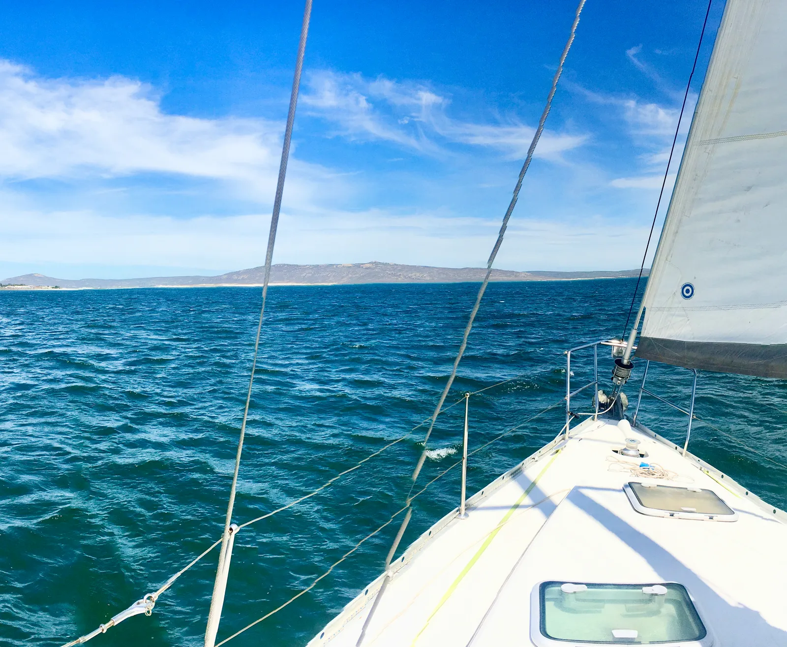 Segelboot mit entfalteten Segeln auf türkisblauem Meer, im Hintergrund kahle Insel unter klarem Himmel